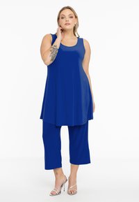 Koningblauwe mouwloze tuniek met een A-lijn snit, gecombineerd met cropped broeken. Gladde stoftextuur, gedragen met zilveren met strikjes.