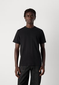 Jonge man met korte dreadlocks die een eenvoudig zwart T-shirt en donkere jeans draagt, staand tegen een effen lichte achtergrond.