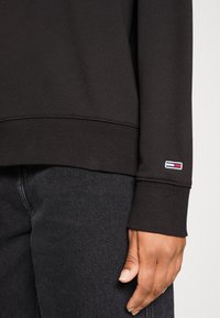 Felpa nera con polsini e orlo a coste, con un piccolo patch logo in rosso, bianco e blu sulla manica. Abbinata a jeans scuri.