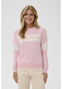 Mujer sonriente con cabello largo y rubio que lleva un suéter de manga larga rosa y blanco con el texto "après tennis" y pantalones beige.