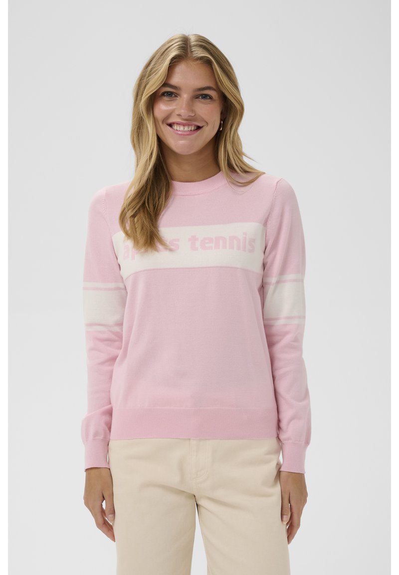 Mujer sonriente con cabello largo y rubio que lleva un suéter de manga larga rosa y blanco con el texto "après tennis" y pantalones beige.
