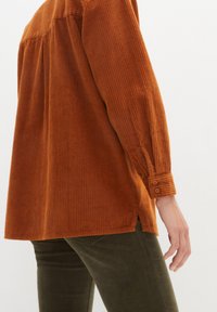 Chemise en corduroy couleur rouille avec des manches longues et des poignets boutonnés ; présente des côtes verticales et des fentes sur les côtés pour une touche de détail supplémentaire.