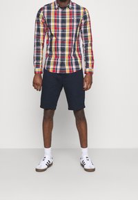Uomo che indossa una camicia a maniche lunghe a scacchi multicolori, pantaloni corti blu, calze bianche e scarpe da ginnastica bianche con strisce nere, in piedi su un pavimento grigio.