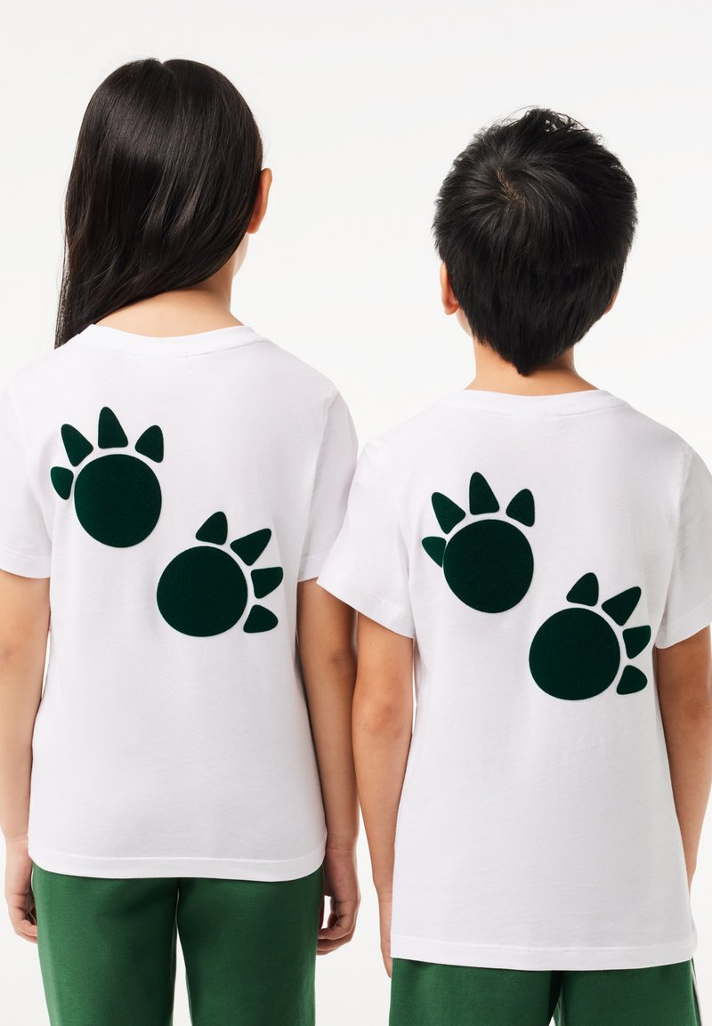 T-shirts blancs avec des motifs de pattes vertes au dos. Manches courtes ; le tissu semble lisse et léger.