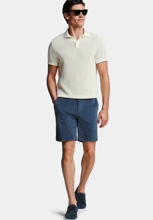 Uomo con occhiali da sole neri, polo a maniche corte in maglia bianca, pantaloncini blu e scarpe stringate blu navy che cammina con una mano in tasca.
