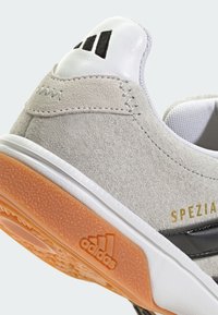 Svetlosivé semišové tenisky s bielou gumovou podrážkou, oranžovým trakčným podošvou a čiernymi akcentmi. Obsahuje zlatý textový logo "SPEZIA".