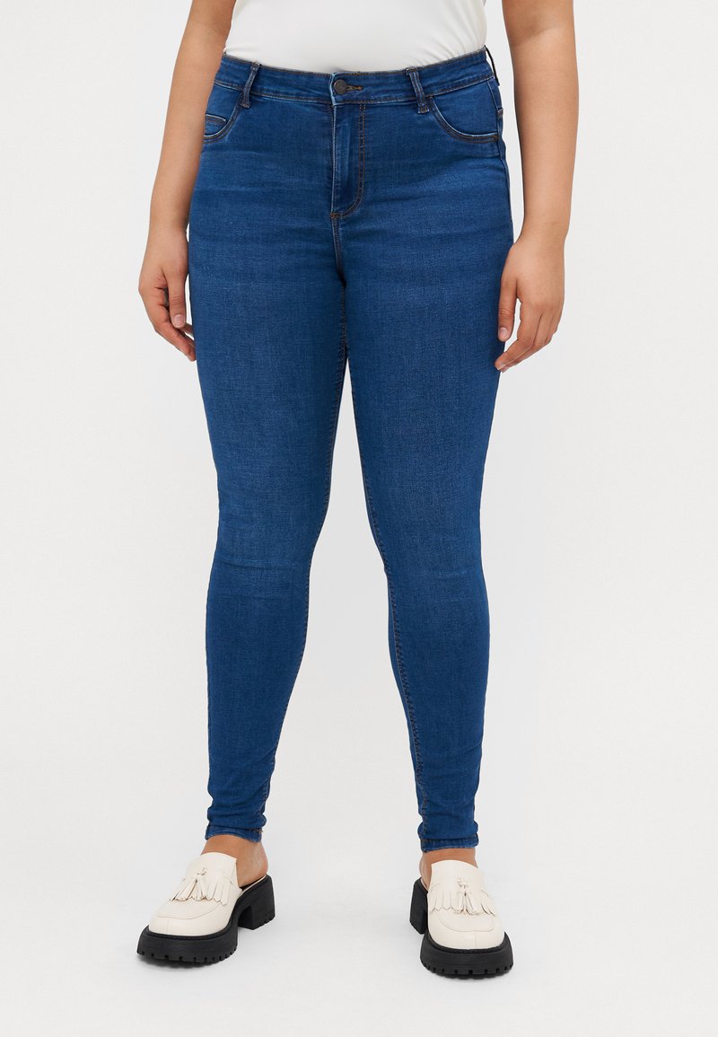 Blue denim skinny jeans with a high waist and subtle fading, paired with white moccasin-style shoes featuring fringe accents.