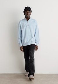 Suéter azul claro con media cremallera, puños acanalados, combinado con jeans oscuros y zapatillas grises; modelo lleva una gorra negra. Diseño sencillo y casual.