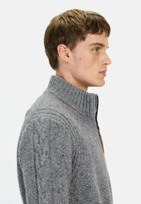 Grijze gebreide trui met een hoge kraag, rits en textuurpatronen. De stof heeft een gespikkeld ontwerp met uitgesproken kabelgebreide accenten.