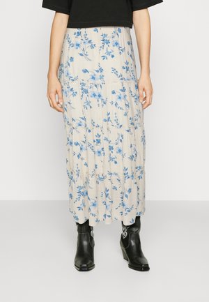 Woman wearing a black top, cream maxi skirt with blue floral print, and black ankle cowboy boots, standing against a plain background.