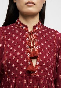 Femme portant une robe bordeaux avec de petits motifs floraux et un col à pampilles nouées orné de broderies.
