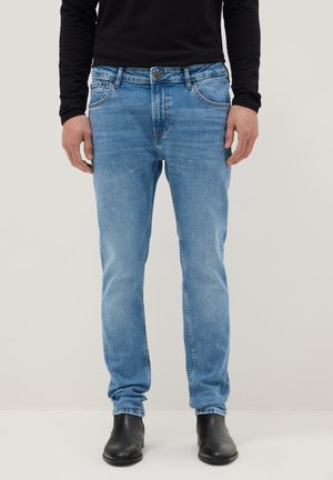 Hombre con jeans ajustados azul claro, camisa de manga larga negra y botines negros, de pie frente a un fondo liso.