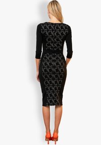 Schwarzes Kleid mit Dreiviertelärmeln, das ein strukturiertes Muster in Silber aufweist. Figurbetonte Silhouette, knielang, kombiniert mit leuchtend orangefarbenen High Heels.