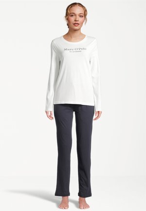 Kobieta stojąca boso, ubrana w białą, długorękawową koszulę Marc O'Polo i ciemne spodnie o prostych nogawkach na jednolitym białym tle.