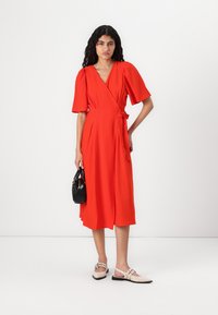 Rochie roșie, cu mâneci scurte și pufoase, de lungime medie, până la jumătatea gambei, legată în talie. Modelul ține o geantă neagră și poartă încălțăminte de culoare deschisă.