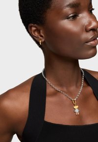 Collar de cadena de plata que presenta un colgante multicolor que se asemeja a una figura de dibujos animados con un acento dorado, llevado con una blusa negra.