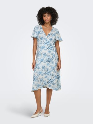 Femme aux cheveux bouclés portant une robe portefeuille fleurie blanche et bleue et des talons slingback blancs, debout devant un fond clair uni.