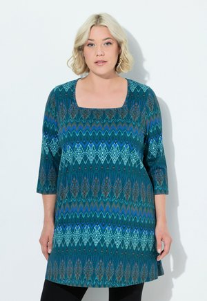 Teal tuniek met een vierkante halslijn, ¾ mouwen en ingewikkelde geometrische patronen in blauwe en bruine tinten. Zachte stof met een relaxte pasvorm.
