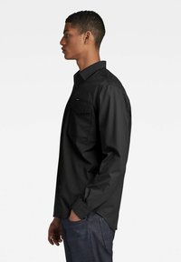 Camisa negra de manga larga con collar abotonado, bolsillo delantero y corte recto. Tejido suave y ligero. Llevada con jeans azules.