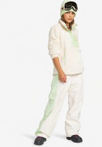 Fleece-Pullover in Creme mit mintgrünen Akzenten; kombiniert mit cremefarbenen Hosen mit mintgrünen Seitenstreifen. Schwarze Schneestiefel und Skibrille.