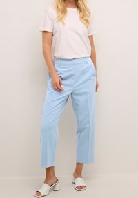 Donna che indossa una camicia bianca a maniche corte, pantaloni cropped azzurri chiari con tasche e sandali con tacco aperti bianchi, in piedi di fronte a un muro neutro.