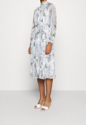 Femme portant une robe midi plissée à manches longues et col haut, avec un imprimé floral pâle et une taille nouée, associée à des talons blancs ouverts à l'avant.