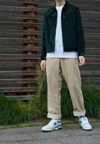 Giacca di velluto a coste verde, T-shirt bianca, pantaloni di velluto a coste beige, sneakers bianche e verdi. Sullo sfondo si vedono listelli di legno e erba.