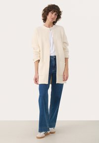 Cream cardigan with textured knit, open front design, and two front pockets, paired with blue jeans and white sneakers.