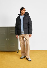 Schwarze Pufferjacke mit Kapuze, mit Reißverschluss und elastischen Bündchen, getragen über einem hellblauen Hemd, kombiniert mit hellbeigen Hosen und grauen Sneakern.