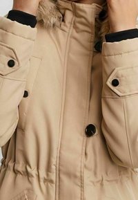 Parka color beige con cappuccio foderato in pelliccia, bottoni a pressione e cintura elastica in vita. Presenta dettagli neri sui polsini e sulle tasche.