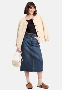 Donna sorridente che indossa una giacca crema con bottoni, un top nero, una gonna midi in denim con cintura, calzini beige e scarpe slip-on marroni e pelose, mentre tiene in mano una borsa crema.