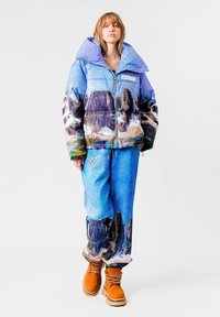 Chaqueta tipo puffa con un estampado de montaña en tonos azules, combinada con pantalones azules texturizados y botas color marrón claro. La chaqueta tiene un gran cuello.