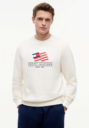 Giovane uomo con felpa bianca Tommy Hilfiger con logo rosso, bianco e blu e pantaloni blu scuro, in piedi con le mani in tasca davanti a uno sfondo bianco.