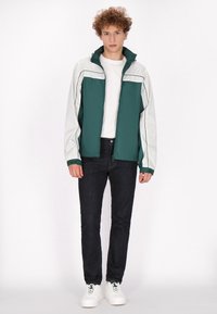 Grön och vit zip-up jacka med hög krage, slät textur och kontrasterande sömmar, bärs över en vit t-shirt och mörka jeans.