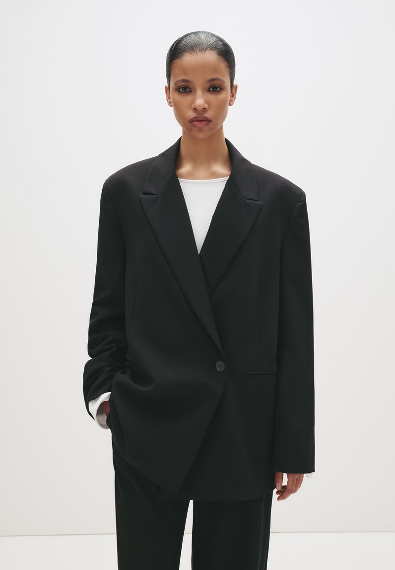 Giovane donna con capelli slicked-back indossa un blazer nero oversize e pantaloni neri sopra una camicia bianca, in piedi contro uno sfondo bianco semplice.