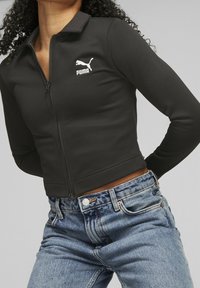 Chaqueta corta con cremallera negra hecha de tela texturizada, con cuello y un logo blanco de Puma en el lado izquierdo del pecho. Combinada con jeans de mezclilla azul.