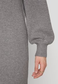 Grå stickad tröja med ribbat mönster, långa ärmar med puffiga axlar och åtsittande ärmslut. En hand med en guldring syns.