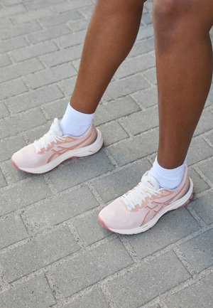 Jambes à la peau brune portant des chaussettes blanches et des chaussures de course rose clair, debout sur un sol pavé gris.