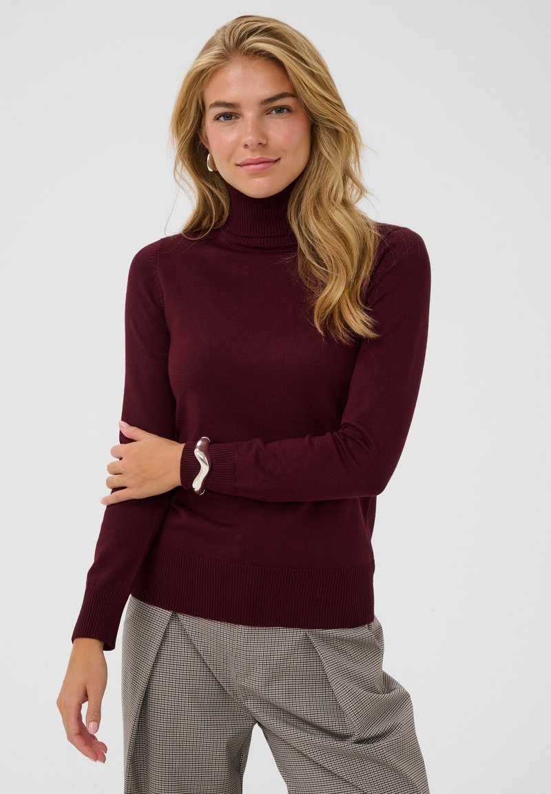Femme aux cheveux blonds ondulés portant un col roulé bordeaux, un pantalon à carreaux, un bracelet en argent et des boucles d'oreilles créoles, debout devant un fond uni.