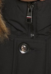 Schwarze Jacke mit einer mit Fell gefütterten Kapuze, ausgestattet mit einem Reißverschluss und einem runden Knopf, der ein Tiergesicht zeigt, beschriftet mit "Hilfiger."