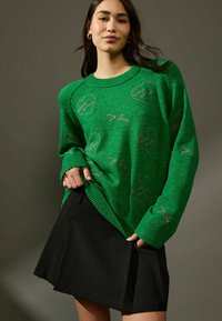 Maglione verde lavorato a maglia con motivi a cuore e testo, colletto e polsini a coste, abbinato a una gonna plissettata nera. Texture liscia, vestibilità casual.