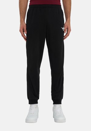 Uomo che indossa pantaloni jogger neri con polsini elastici e sneakers bianche, in piedi davanti a uno sfondo semplice.