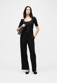 Γυναίκα που στέκεται, ντυμένη με μαύρο τοπ με τετράγωνη λαιμόκοψη, μαύρο παντελόνι με φαρδιά μπατζάκια, μαύρες γόβες και κρατά μια μαύρη τσάντα ώμου.