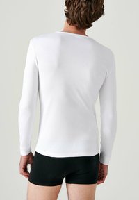 Camicia bianca a maniche lunghe realizzata in morbido tessuto, stile aderente, scollatura rotonda, abbinata a boxer neri, vista da dietro.