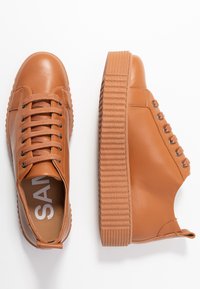 Baskets en cuir tan avec une semelle texturée et striée, un bout rond et des lacets plats. L'intérieur présente un logo de marque visible.