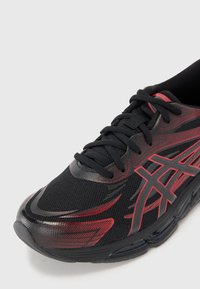 Tênis de corrida preto com uma parte superior texturizada e detalhes em vermelho, além de um logótipo em destaque. O design inclui atacadores e uma sola acolchoada.