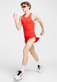 Punane sportlik tank top ja lühikesed püksid, millel on Nike logo. Mudel kannab prille ja valgeid tossusid punaste aktsentidega, joostes ühtlasel taustal.