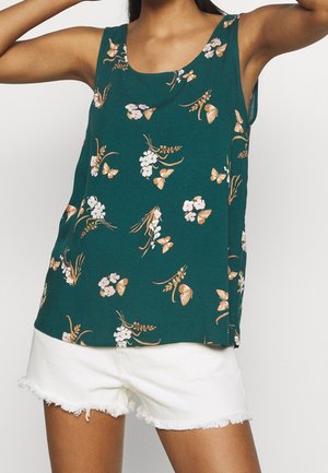 Vrouw met een teal mouwloze top met vlinder- en bloemenprint en witte denim shorts met rafelige randen tegen een witte achtergrond.