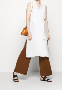 Weißes ärmelloses Kleid mit einem taillierten Gürtel, kombiniert mit braunen weiteren Hosen und schwarzen Sandalen. Enthält eine kleine orange Schultertasche.