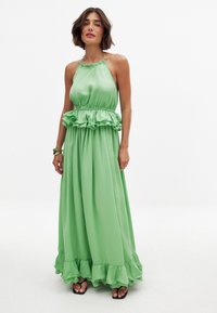 Vihreä maxi-mekko, joka on valmistettu sileästä satiinista, jossa on röyhelövyötärö ja -haalari. Olkaimeton malli, jossa on kerrottu kaula-aukko ja virtaava kangas.